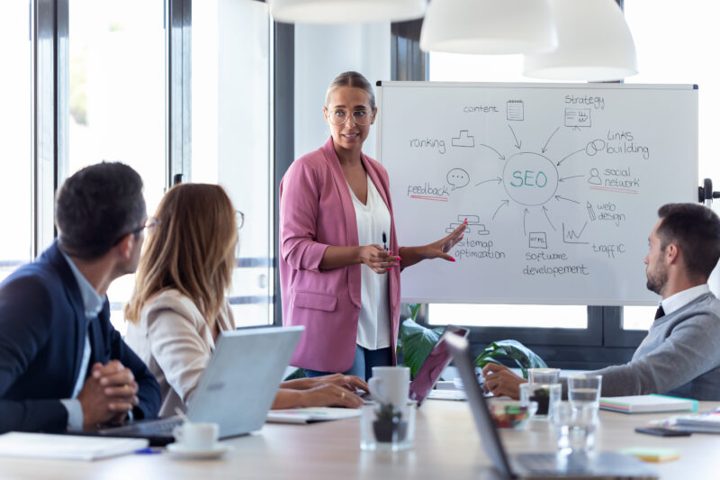 Elegant young businesswoman pointing at white blackboard and exp businesswoman explaining to marketing team how to boost seo for campaigns