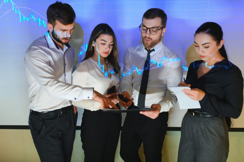 Group of business people working at modern office.Technical price graph, red and blue candlestick chart and stock trading computer screen background. Double exposure. Traders analyzing data AI for sales - sales team using AI tools to boost sales